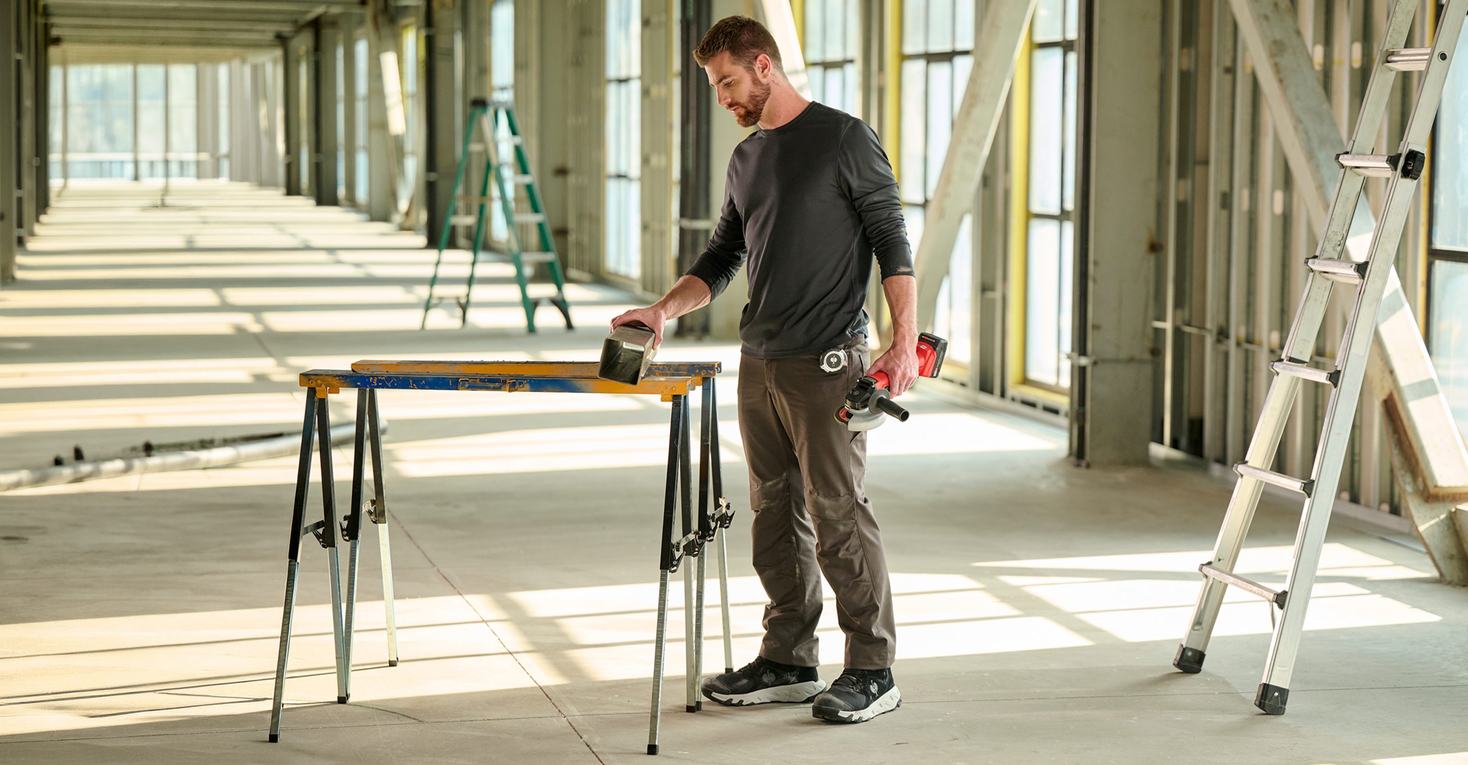 A man in a long-sleeve shirt stands in a large, bright room, holding tools and working at a sawhorse table. Ladders are visible in the background along with large windows.