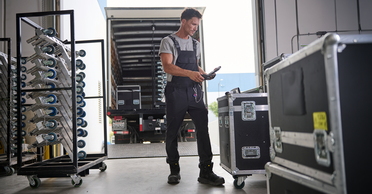 A man in work overalls uses a tablet while standing near large equipment cases in a warehouse, with a truck parked at a loading dock in the background.