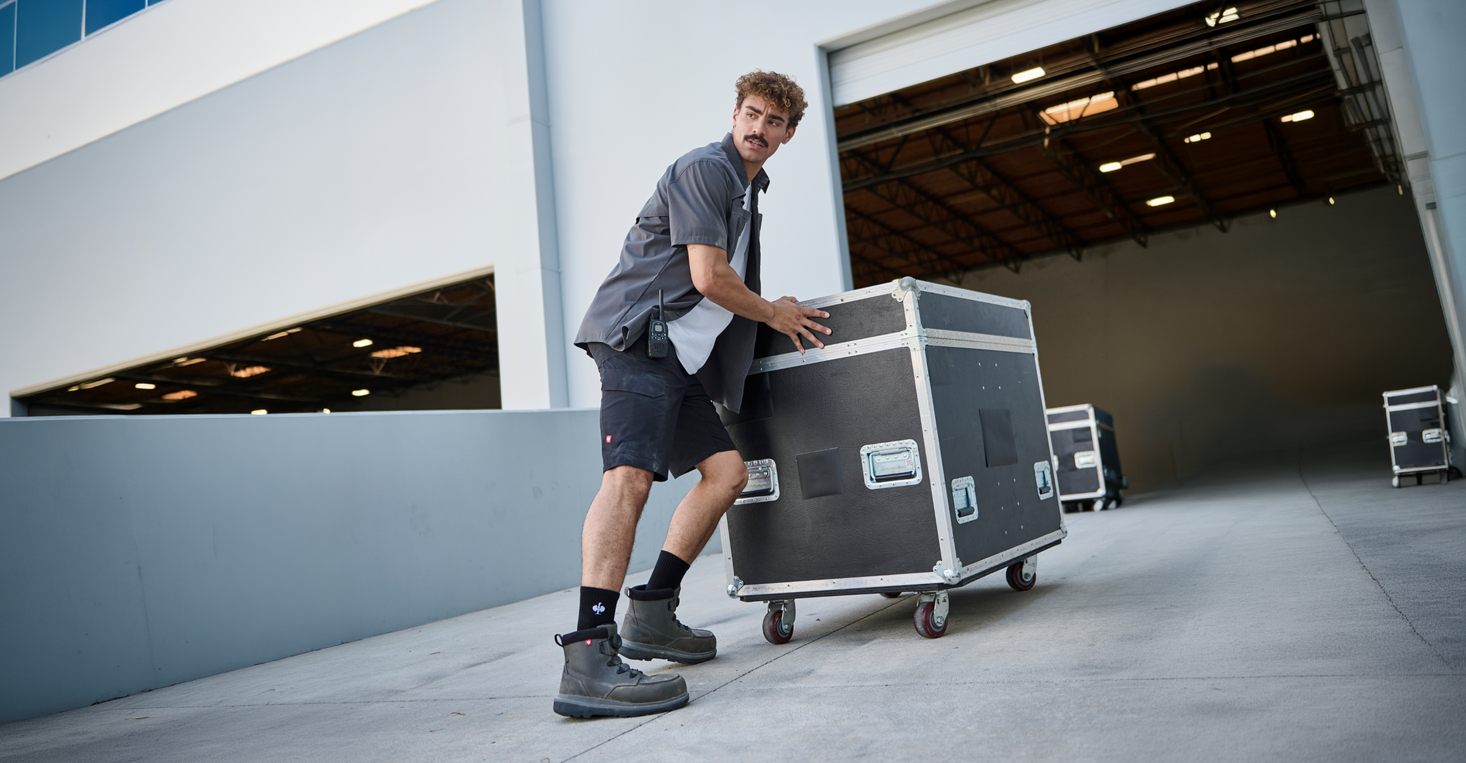 A man in work clothes pushes a large black equipment case on wheels outside an industrial building with an open loading dock door.