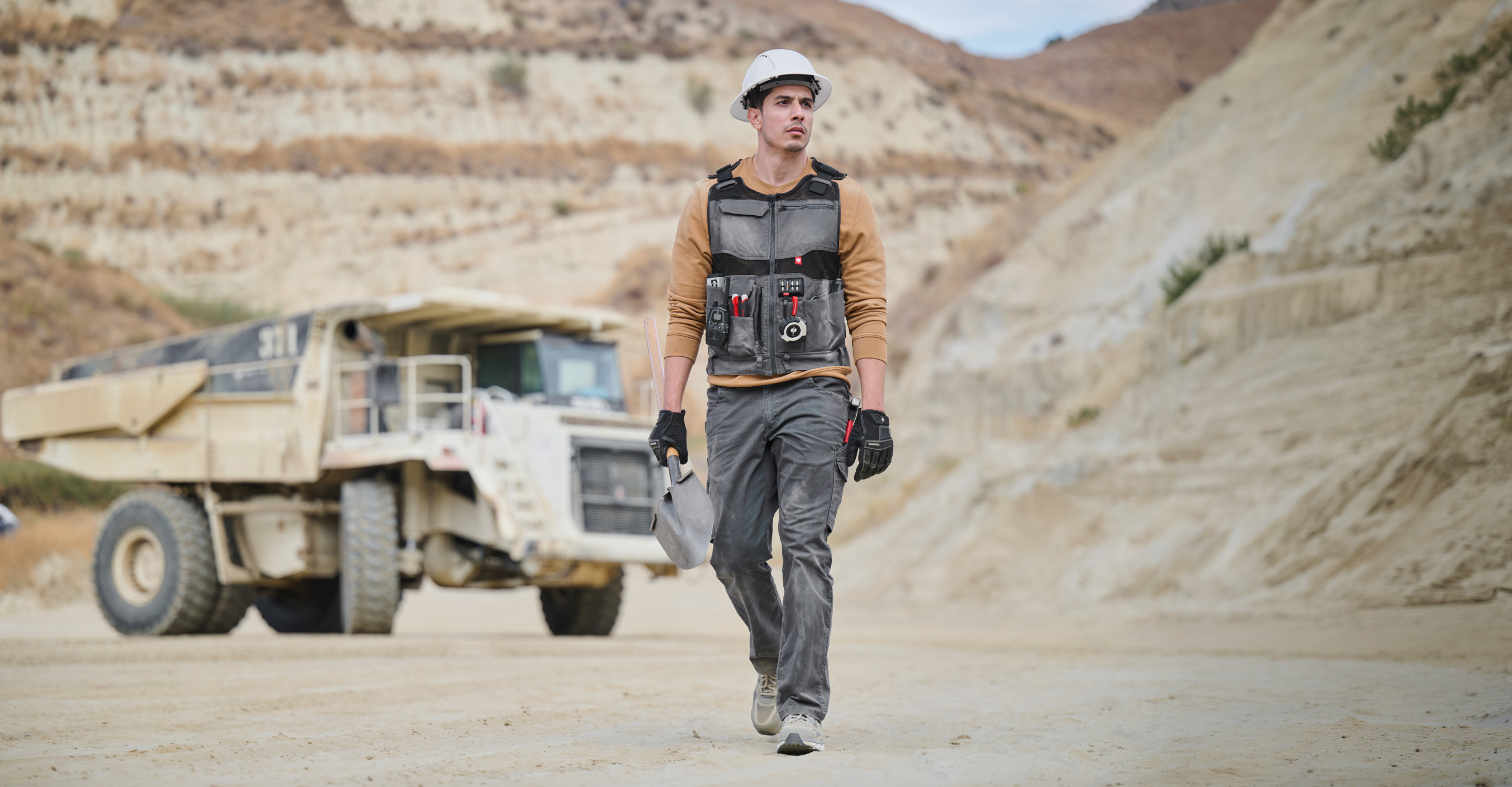A construction worker wearing a white hard hat, a black utility vest over a brown shirt, and gray pants walks in a dusty quarry with a large dump truck in the background.