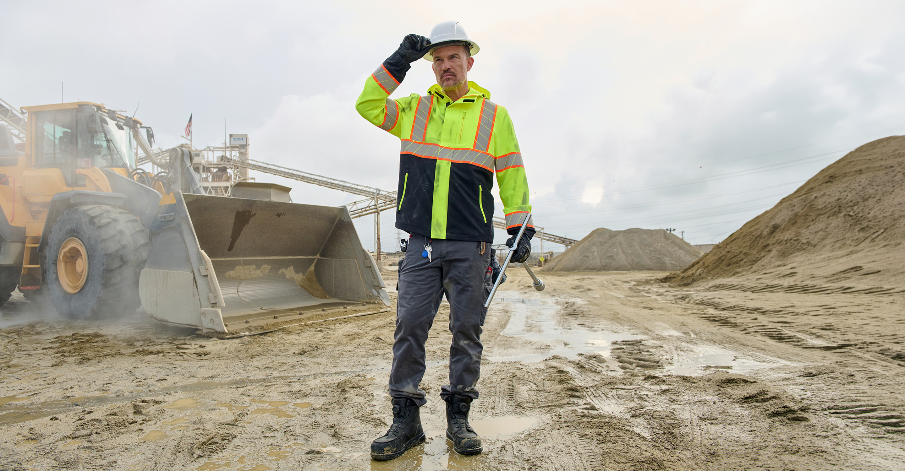A construction worker wearing a high-visibility neon yellow and navy jacket, gray pants, black boots, and a white hard hat stands in a muddy outdoor site next to a yellow front-end loader under an overcast sky.