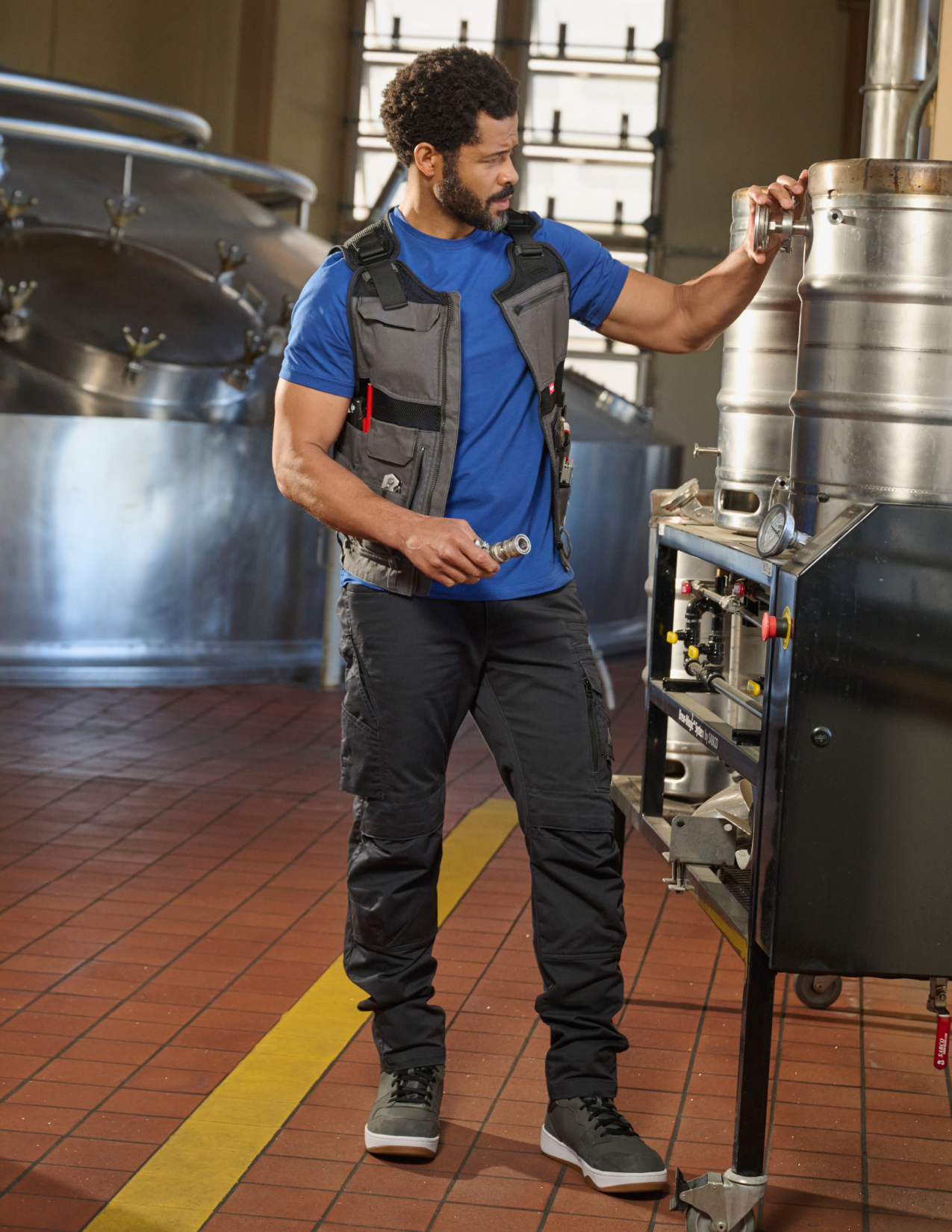 Person in a brewery setting with large metal tanks and a tool vest.